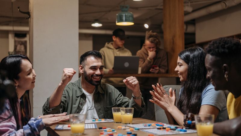 Friends around a table mid board game, one of them celebrating a turn.