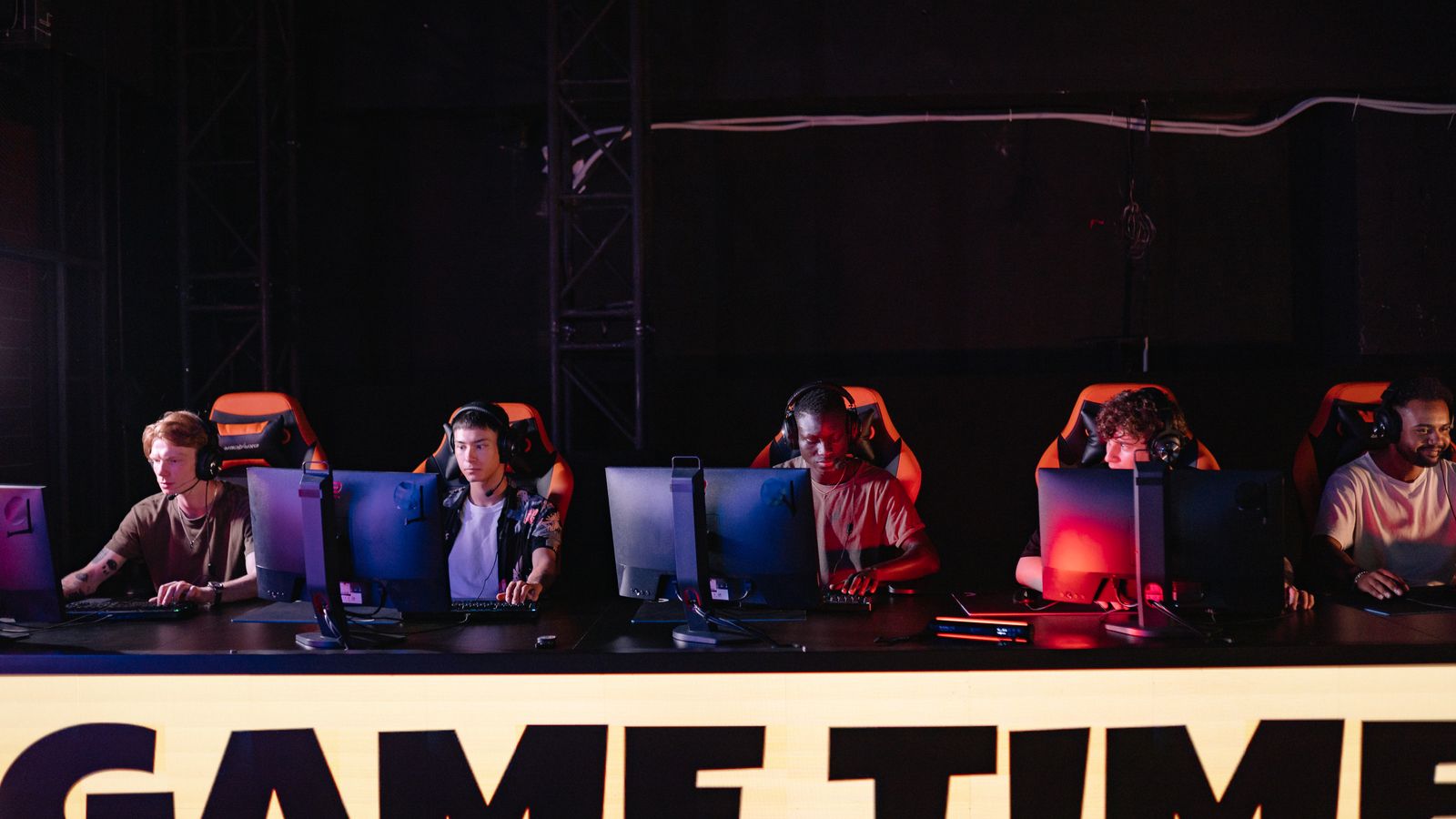 A row of players at a lit tournament stage, monitors facing away, under a GAME TIME banner.